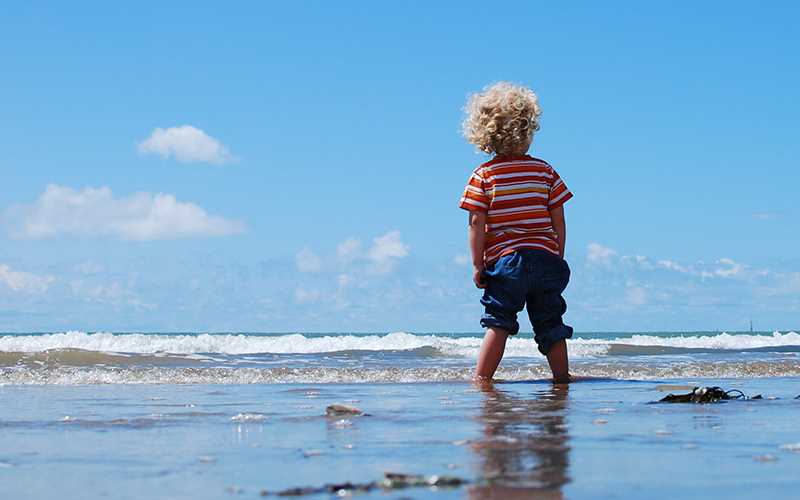Et lite barn står med føttene i havet på en strand.